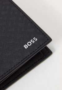 Juodas tekstūruotas piniginė su subtiliu reljefiniu raštu. Priekinėje pusėje yra sidabrinė „BOSS“ logo, su siuvinėtomis briaunomis ir stilingu dizainu.