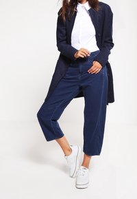 Pantaloni a gamba larga blu navy con vita alta, abbinati a una camicia bianca con bottoni indossata sotto un cappotto scuro. Indossati con sneaker bianche.