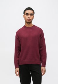 Bordowy sweter z dzianiny z okrągłym dekoltem, ściągaczami na rękawach i u dołu. Charakteryzuje się luźnym krojem i fakturowaną tkaniną. W połączeniu z czarnymi spodniami.