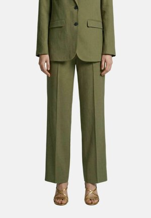 Persona con pantalones y chaqueta de traje verde oliva que está de pie con las manos relajadas a los lados y sandalias doradas con tiras y tacón alto.