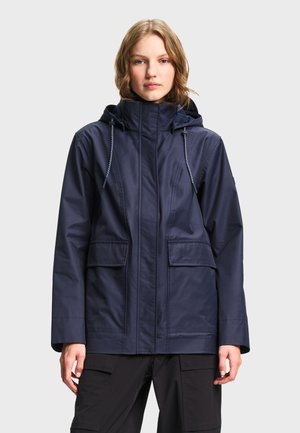 Mujer con chaqueta impermeable azul marino con capucha, bolsillos delanteros y cordones, de pie frente a un fondo claro y liso.