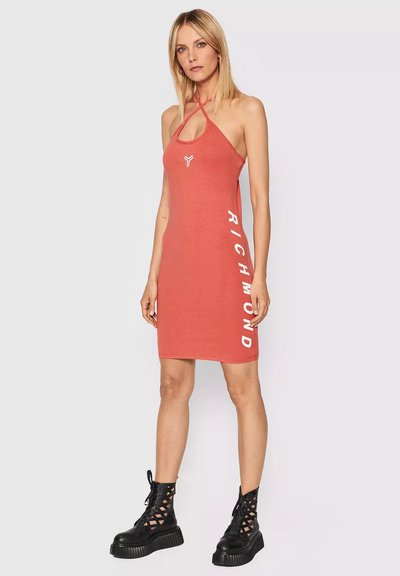 Donna bionda che indossa un mini abito senza maniche color corallo con la scritta "RICHMOND" e stivali platform neri a rete, in piedi su uno sfondo neutro.