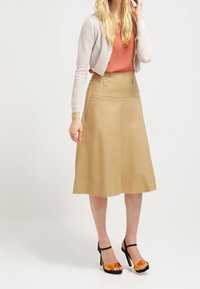 Žena ve korálovém topu, béžovém zkráceném kardiganu, tan midi sukni ve tvaru A a černých sandálech na vysokém podpatku s oranžovými pásky.