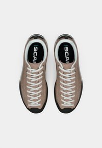Sapatos de camurça castanho claro com atacadores brancos e sola de borracha preta, apresentando uma palmilha texturada preta e acentos de marca discretos.