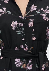 Μαύρο φόρεμα με ροζ και πράσινο floral μοτίβο, υφασμάτινα κουμπιά μπροστά, τσέπη στο στήθος και ζωνάκι δεμένο στη μέση.