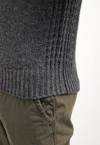 Gros plan d'une personne portant un pull en maille gris foncé et un pantalon vert olive, montrant la texture et les détails du tissu.