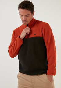 Fleece pullover with an orange top and black bottom, half-zip closure, and small logo detail on the chest; features a relaxed fit.