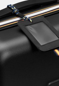 Valigia rigida nera con finitura lucida, dotata di una zip con superficie testurizzata e di un porta etichetta trasparente. Accentuata da una tracolla intrecciata multicolore.