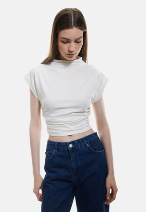 White cropped top with short sleeves and a high neckline, featuring a gathered texture at the waist, paired with dark blue high-waisted jeans.