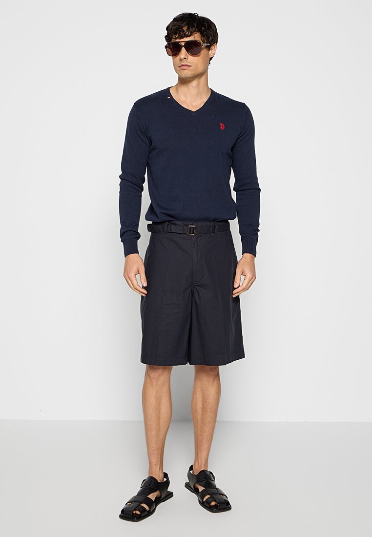 Marineblauer V-Ausschnitt-Pullover, schwarze Shorts mit Gürtel und schwarze Sandalen. Modell trägt Sonnenbrille und steht vor einem neutralen Hintergrund.