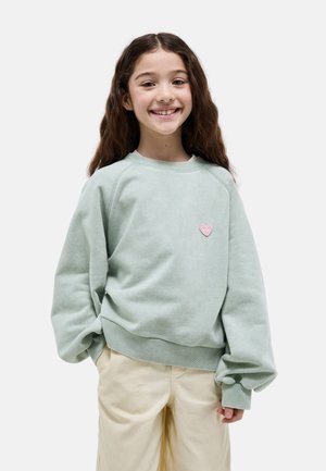 Fille souriante aux longs cheveux bruns portant un sweat-shirt ample vert clair avec un petit cœur rose sur le côté gauche de la poitrine et un pantalon beige.