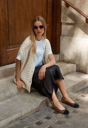 Naine pikkade blondide juustega, päikeseprillid, valge särk, kootud vest, must seelik ja pruunid madalad kingad, istub kivist trepil puuuks e ees.