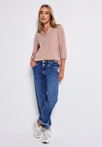 Chemise henley rose clair à manches longues avec trois boutons, associée à un jean bleu à coupe ample. Des baskets blanches complètent la tenue.