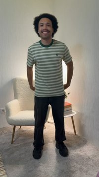 Bărbat zâmbitor cu păr creț, purtând un tricou în dungi și pantaloni negri, stă într-o cameră cu mochetă, lângă un fotoliu alb și o măsuță rotundă mică cu cărți.