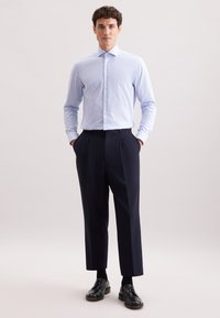 Homme debout portant une chemise à rayures bleu clair et blanche à manches longues, un pantalon plissé sombre, des chaussettes noires et des chaussures habillées noires sur un fond uni.