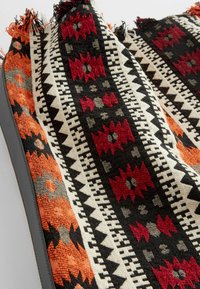 Gewebter Stoff mit geometrischen Mustern in Rot, Orange, Schwarz und Weiß. Strukturierten Oberfläche mit Fransenrändern und einfarbigem grauen Besatz.