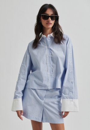 Camicia oversize a righe blu e bianche con colletto bianco e polsini abbottonati, abbinata a pantaloni corto coordinati. Tessuto morbido e vestibilità comoda.