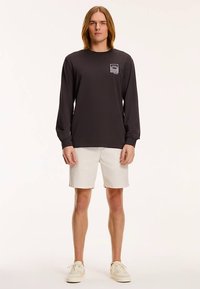 Czarna koszulka z długim rękawem z grafiką na piersi, zestawiona z białymi szortami i jasnymi trampkami. Model stoi prosto, zwrócony przodem.