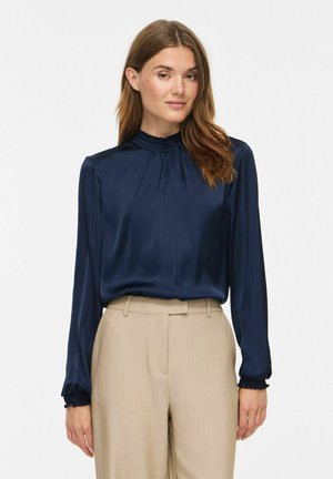 Femme portant un chemisier bleu marine à manches longues avec des poignets volantés et un pantalon beige taille haute, debout devant un fond blanc.