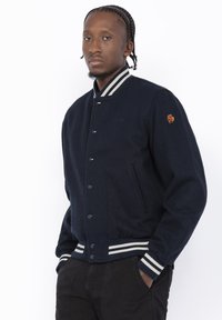 Man met gevlochten haar, gekleed in een marineblauwe varsityjack met witte gestreepte kraag, manchetten en zoom, staande met de handen in de zakken.