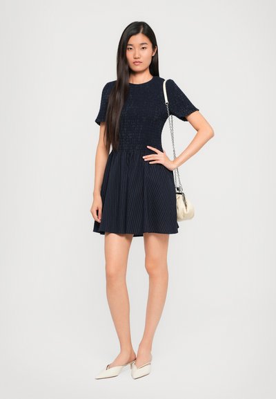 Tengeri ruházat rövid ujjakkal és redőzött szoknyával, függőleges csíkokkal és testhez simuló felsőrésszel. Kiegészítve egy kis krémszínű válltáskával és mules lábbelivel.