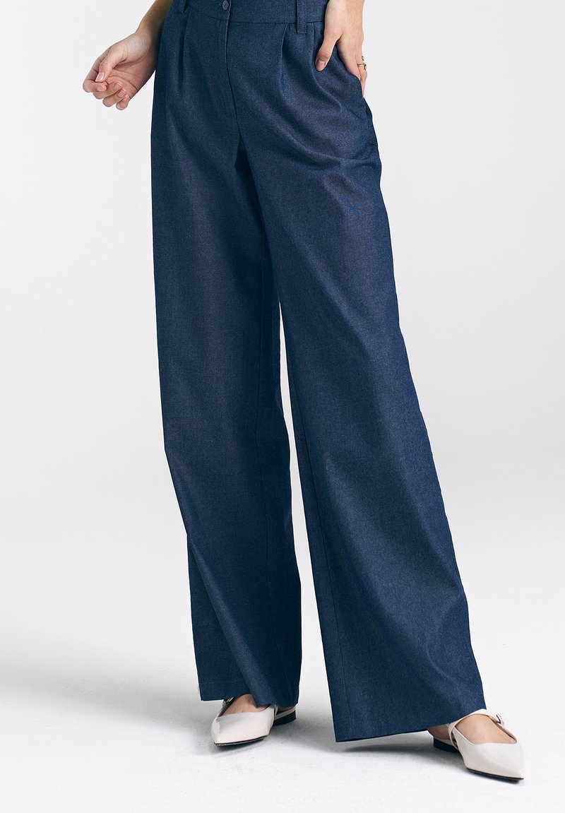 Spodnie o szerokiej nogawce z ciemnego denimu, o dopasowanym wyglądzie, z przyszytymi fałdami z przodu i bocznymi kieszeniami. W zestawie z jasnymi butami wsuwanymi.