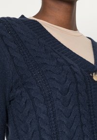 Marinblå kabelstickad cardigan med djup V-hals, som har ett framträdande texturerat mönster, ribbade kanter och en enda rund knappstängning.