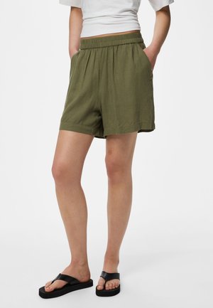 Kvinde iført olivengrønne elastiske shorts i taljen, hvid cropped T-shirt og sorte platformssandaler, står med hænderne i lommerne.