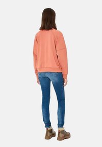 Un sweat à capuche rose avec une coupe décontractée, des poignets et une taille côtelés, associé à un jean bleu et des bottines brunes. La vue montre l'arrière.