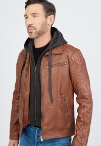 Ruskea nahkatakki, jonka alla on harmaa huppari, jossa on vetoketjulliset taskut ja istuva malli. Sujuva rakenne ja ribbimäiset yksityiskohdat.