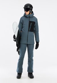 Snowboarder outfit med en blå-grå vandtæt jakke med en sort brystlomme, matchende bukser, handsker og en sort hjelm med goggles.