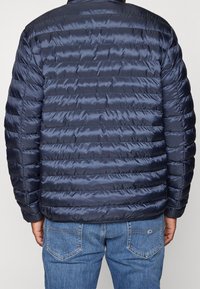 Granatowa kurtka puchowa z poziomymi przeszyciami, gładką tkaniną i luźnym krojem. Zestawiona z jasnoniebieskimi dżinsami.