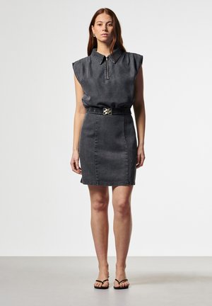 Vrouw draagt een mouwloze zwarte spijkerjurk met een ritssluiting bij de kraag en een ceintuur in de taille, staand voor een effen lichtgrijze achtergrond.