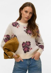 Balts teksturēts džemperis ar violetiem un rozā ziedu rakstiem, apaļu kakla izgriezumu un garām piedurknēm, rokās tur mostardas krāsas somu.