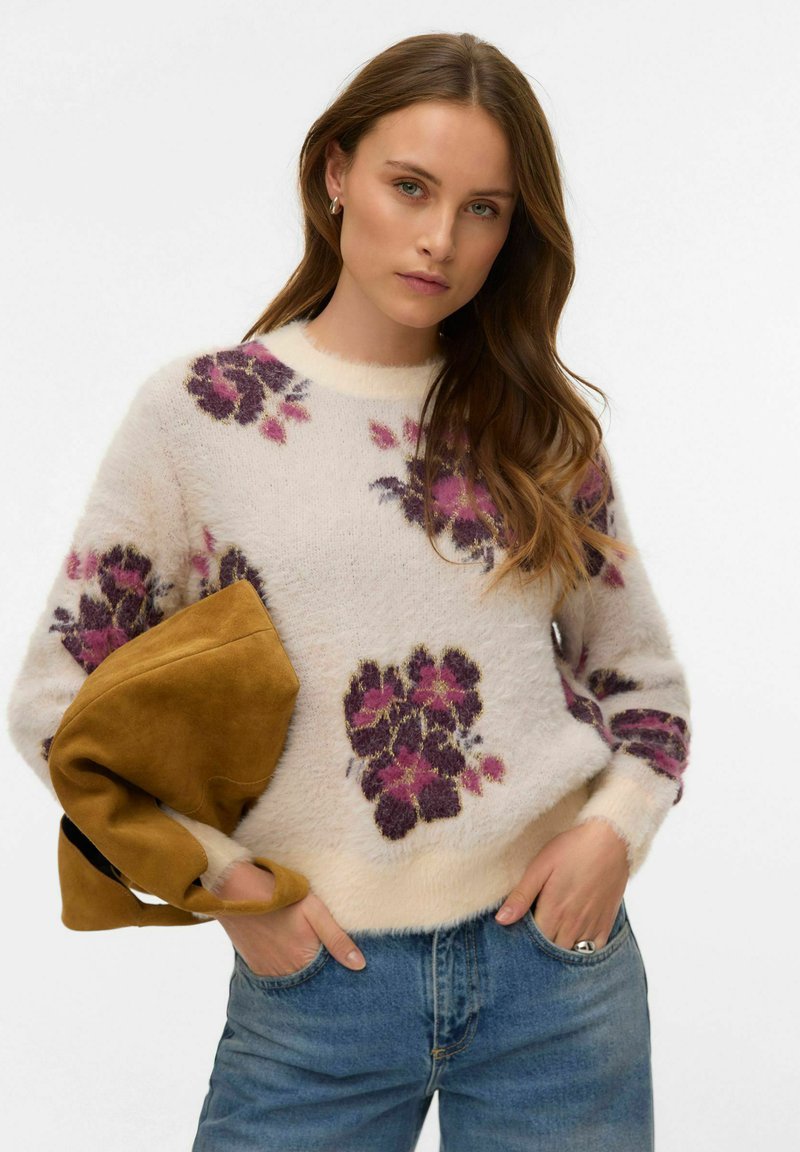 Balts teksturēts džemperis ar violetiem un rozā ziedu rakstiem, apaļu kakla izgriezumu un garām piedurknēm, rokās tur mostardas krāsas somu.