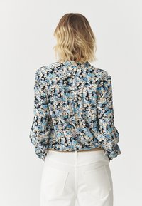 Květinová blůza v modré, černé a krémové barvě s límcem na knoflíky, dlouhými rukávy a nařasenými manžetami, zobrazená zezadu s bílými kalhotami.