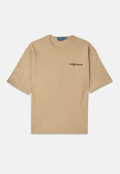 Beige kokvilnas T-krekls ar īsām piedurknēm, apaļu dekoltē un mazu melnu logo kreisajā krūtīs. Mīksta tekstūra.