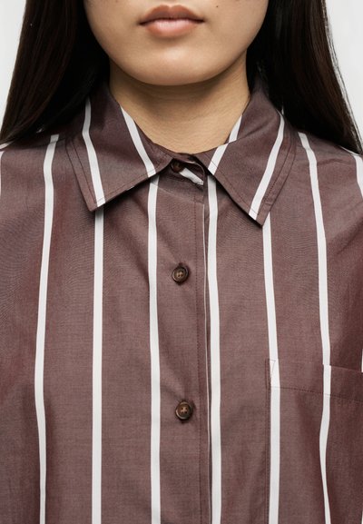 Κοντινό πλάνο ενός ατόμου που φοράει καφέ πουκάμισο με κάθετες λευκές ρίγες και σκουρόχρωμα κουμπιά, εμφανίζοντας το γιακά και το άνω μέρος του στήθους.