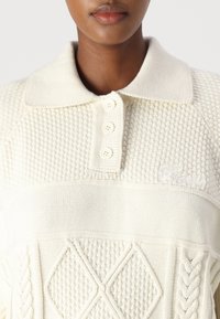 Weißes gestricktes Poloshirt mit einem strukturierten Muster, drei Knöpfen und einem Rautenmuster aus Kabelstrick auf der Vorderseite. Dezentes Markenlogo auf der Brust.