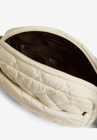 Borsa a tracolla di colore crema trapuntata con apertura zip. L'interno è foderato in tessuto scuro e presenta una piccola tasca con zip.