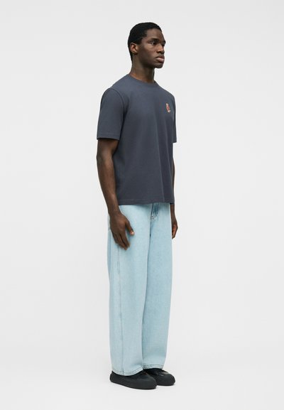 Άνδρας που στέκεται φορώντας σκούρο γκρι κοντομάνικο μπλουζάκι με μικρό λογότυπο και ανοιχτό μπλε jeans με φαρδιά μπάρα και μαύρα παπούτσια.