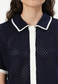 Polo top in maglia blu navy con colletto bianco e pattina dei bottoni; presenta una texture a trama aperta e maniche corte.