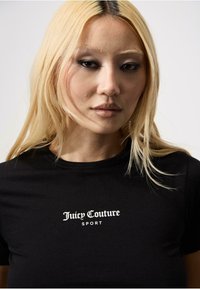 Juodos medvilnės marškinėliai su "Juicy Couture SPORT" logotipu baltos spalvos, klasikiniu apvaliu kaklu, minkšta tekstūra ir prigludusia forma.