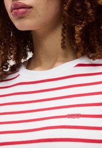 Άτομο με κατσαρά μαλλιά που φοράει λευκό πουκάμισο με οριζόντιες κόκκινες ρίγες και ένα μικρό κόκκινο λογότυπο Levi's στο στήθος.