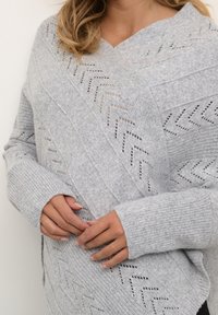 Kvinna med axellångt hår som bär en ljusgrå stickad tröja med ett chevron-mönster, hållande sina händer framför sig.