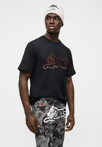 Μαύρο μπλουζάκι με πολύχρωμο σχέδιο από χάντρες, συνδυασμένο με μαύρο παντελόνι με σχέδια που απεικονίζει μια λευκή γάτα και κόκκινες λεπτομέρειες.