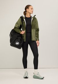 Olijfgroene waterdichte jas met grijze accenten, zwarte leggings, lichtgroene schoenen en een zwarte sporttas met een logo.
