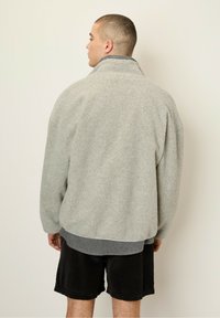 Hellgraue Fleecejacke mit einem hohen Kragen und strukturiertem Oberflächenmaterial. Trägt man über einem dunkelgrauen Shirt und schwarzen Cords-Shorts.