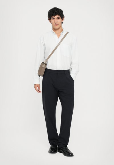 Weiß gestreiftes Hemd mit Knöpfen und Tasche, schwarze Hose und schwarze Schuhe. Eine beige Tasche mit Schulterriemen rundet das Outfit ab.
