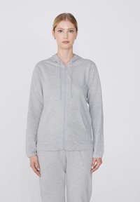 Šedá mikina na zip vyrobená z měkké tkaniny, s kapucí, stahovacími šňůrkami, předními kapsami a úzkými manžetami. Hladká textura, volný střih.